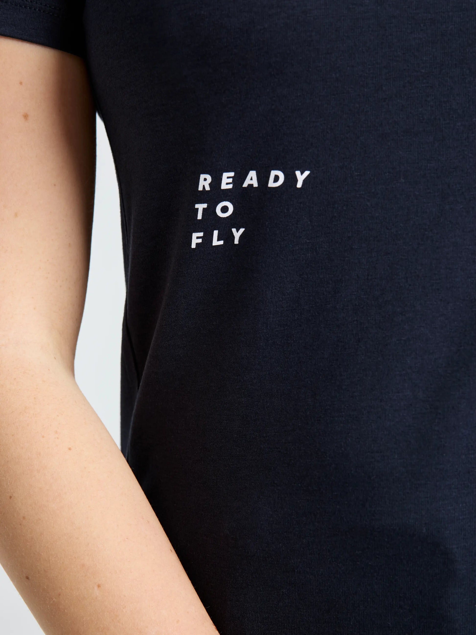 A close-up of a person wearing the SLAM GRAPHIC WS T-SHIRT SS in black, featuring “READY TO FLY” in white uppercase letters on the left chest. The bamboo cotton fabric enhances both comfort and style. Only part of the arm and torso are visible.