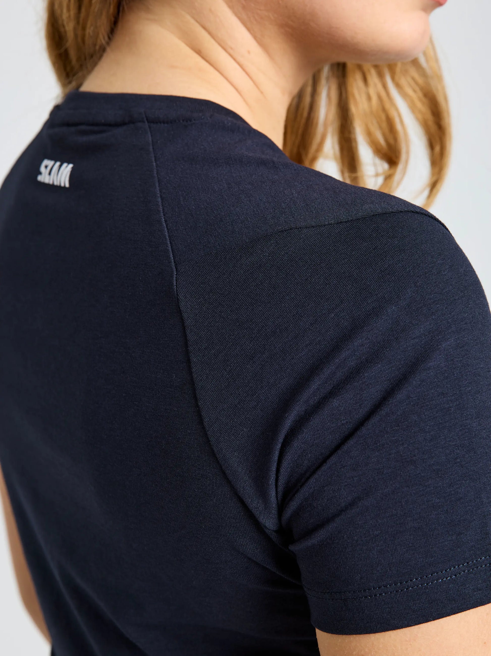 A close-up of a person wearing the SLAM GRAPHIC WS T-SHIRT SS, a navy blue short-sleeve bamboo cotton shirt with small white text near the neckline; their light brown hair is partially visible.