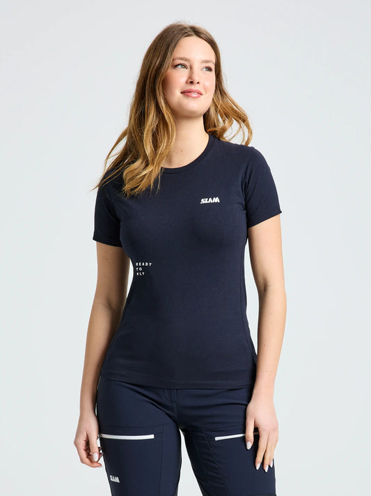 A woman with long light brown hair wears the SLAM GRAPHIC WS T-SHIRT SS, a fitted navy sailing tee, and matching navy pants with white accents. She stands facing forward, slightly smiling with her arms relaxed at her sides.
