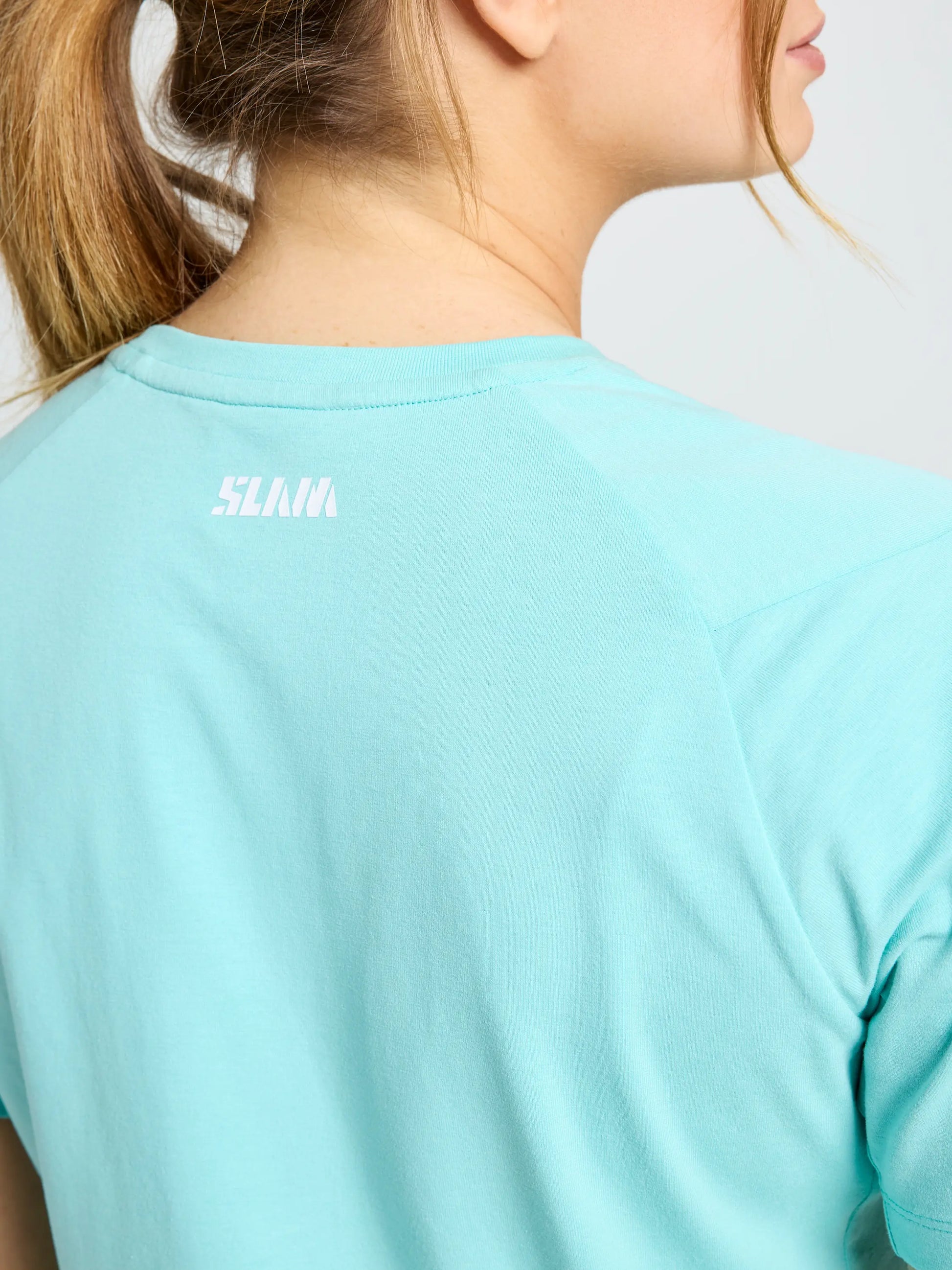 A person with light brown hair in a ponytail is seen from behind, wearing the SLAM GRAPHIC WS T-SHIRT SS in light blue with "SLAM" printed in white on the upper back near the neckline. Part of their profile is visible.