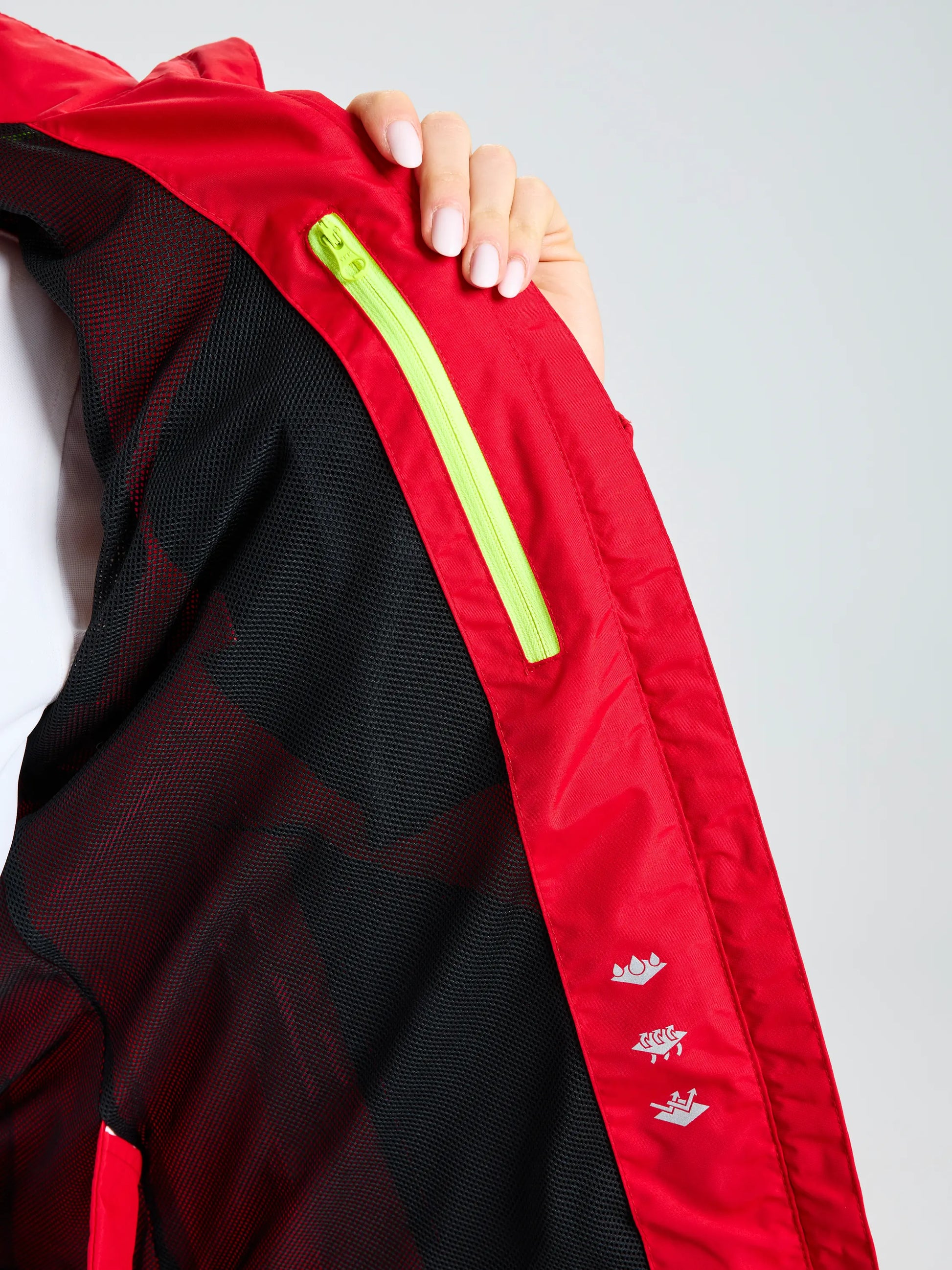 A person holds open the SLAM WS SAILING JACKET, showing its neon green zipper pocket, black mesh lining, and small white icons printed on the inner edge.