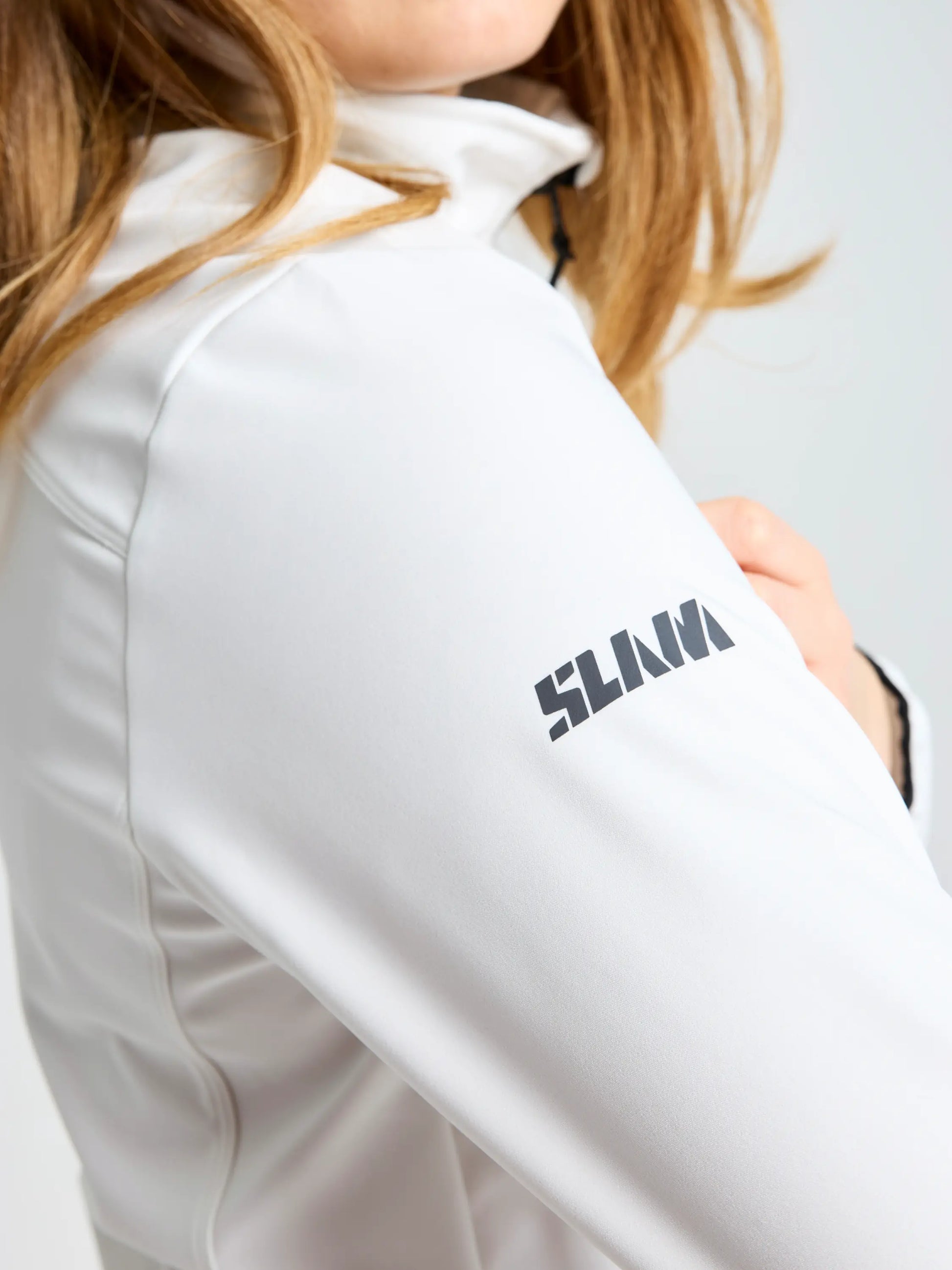 A person with long, light brown hair wears the SLAM ACT WS SOFTSHELL JACKET in white. The sleeve displays a black "SLAM" logo near the shoulder. The background is plain and light-colored.