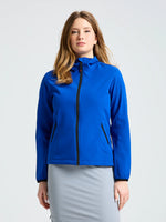 A person with long, light brown hair wears the SLAM ACT WS SOFTSHELL JACKET—a bright blue women’s jacket with black zippers and elastic cuffs—paired with a light gray skirt, standing against a plain light background.