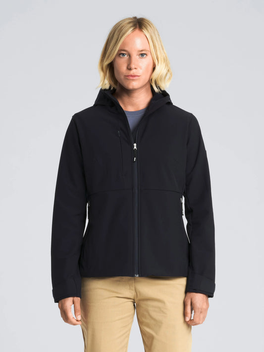 A person with short blonde hair wears the SLAM ALTURA WS WARM SSHELL HOO women’s softshell jacket in DARK NAVY, paired with beige pants and a blue top. They are standing facing forward against a plain light gray background with a neutral facial expression, showcasing the jacket’s thermal comfort suitable for sailing or everyday wear.