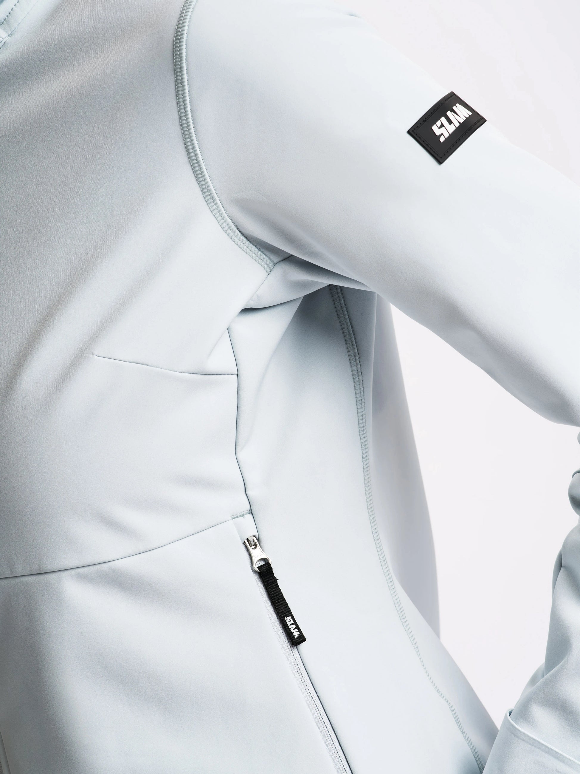 Close-up of the SLAM ALTURA WS WARM SSHELL HOO women’s softshell jacket in light gray, highlighting a zippered pocket, seam details, and a black rectangular ELXN patch on the upper sleeve. The smooth, slightly stretchy fabric is designed for thermal comfort. No background details visible.