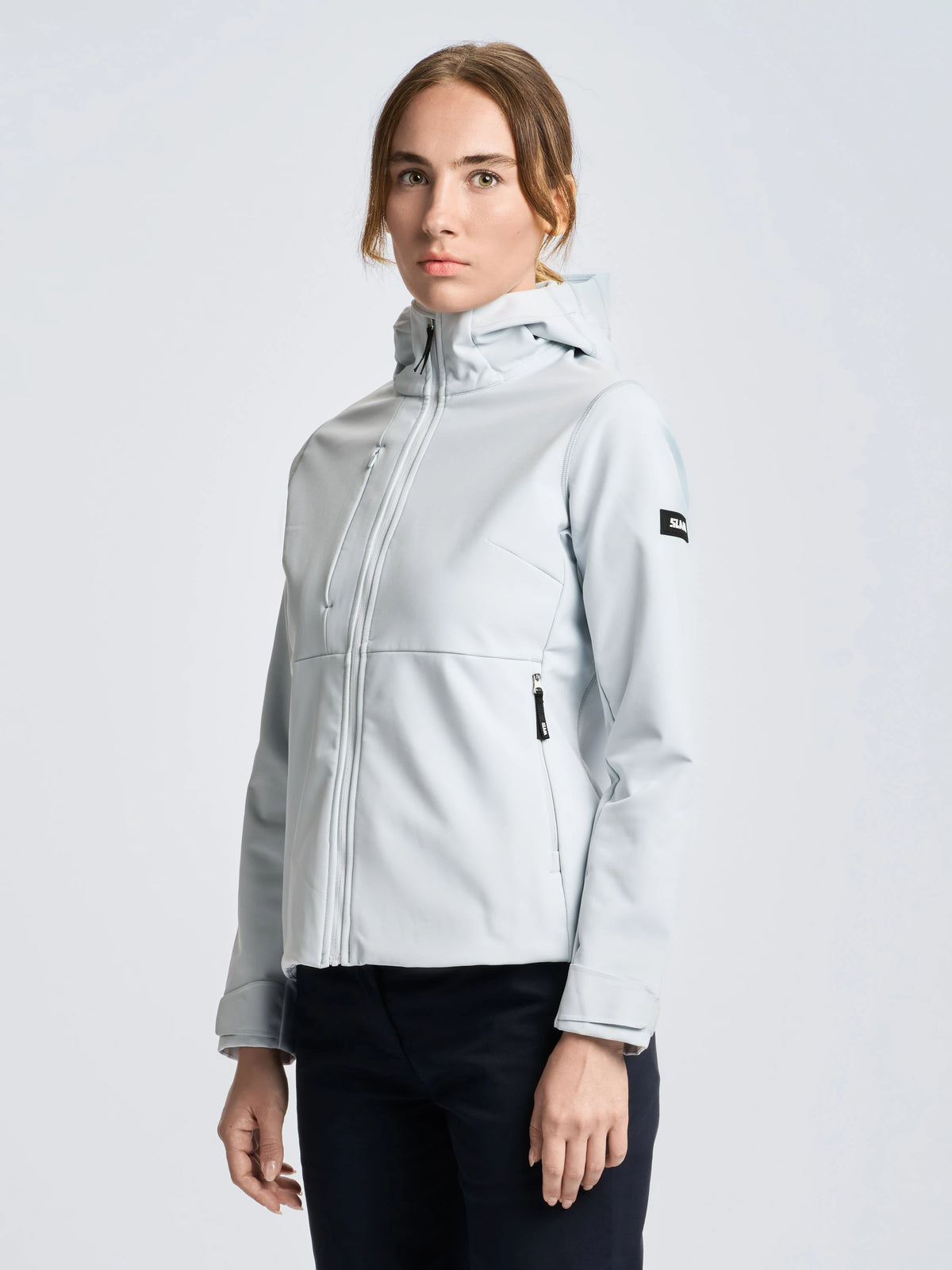 A person stands facing forward against a plain light background, wearing black pants and the SLAM ALTURA WS WARM SSHELL HOO—a light gray women’s softshell jacket with a front zipper. Designed for thermal comfort, the jacket features a black logo patch on the left upper arm.