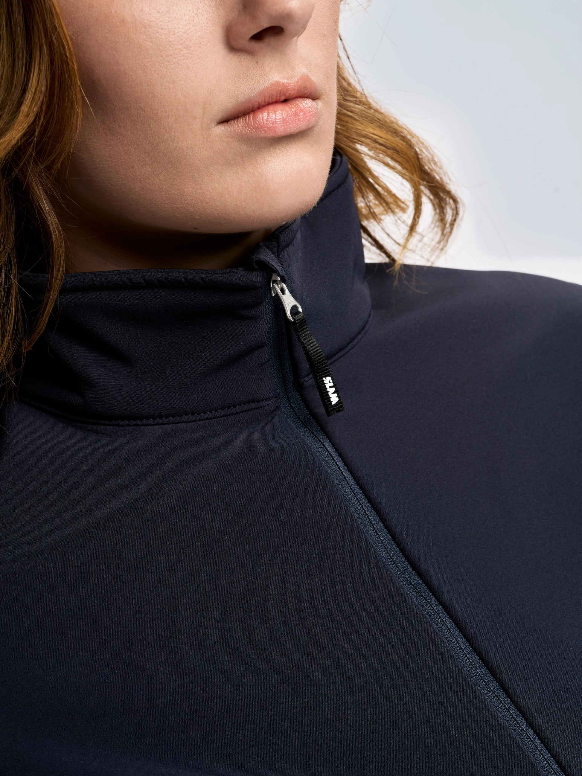 Close-up of a person wearing the SLAM ALTURA WS WARM SSHELL JKT, a dark women’s softshell jacket with the zipper partially closed. Only the lower half of the face, part of the shoulder, and some hair are visible against a neutral, out-of-focus background.