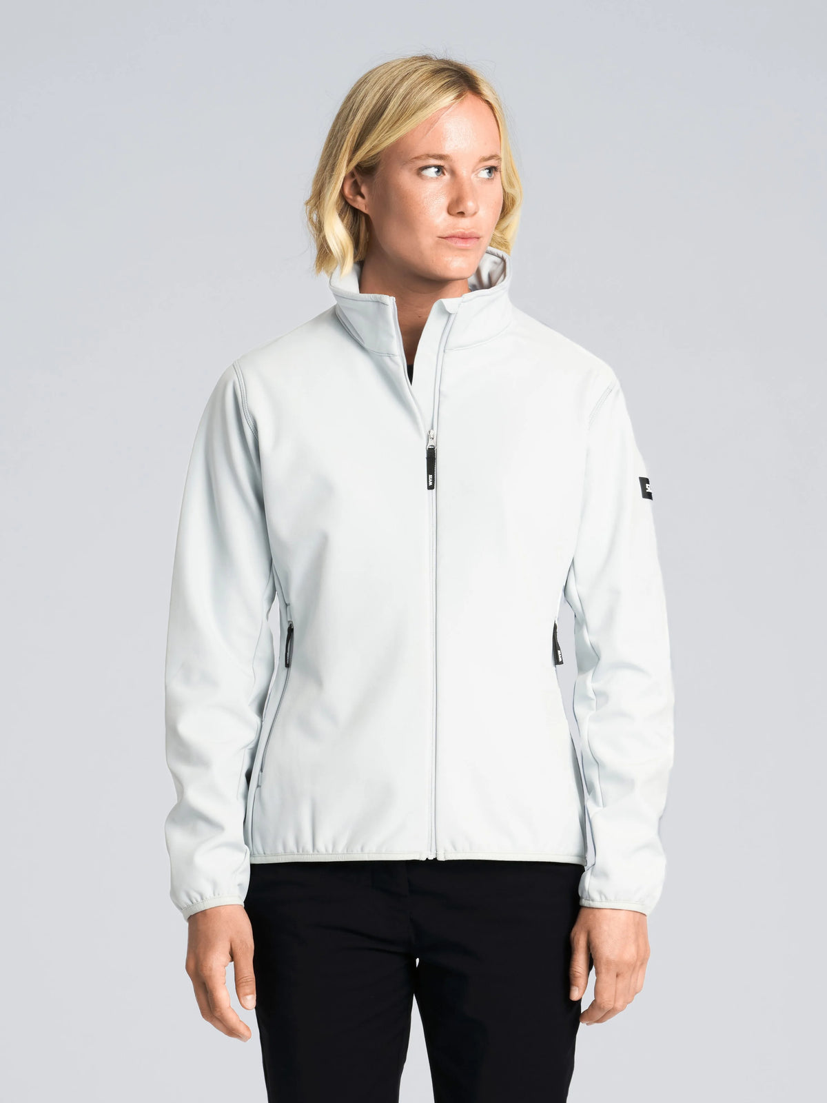 A person with short blond hair is wearing the SLAM ALTURA WS WARM SSHELL JKT in CLIFF GREY, featuring black zippers, along with black pants. They are standing against a plain light gray background and looking slightly to their left.
