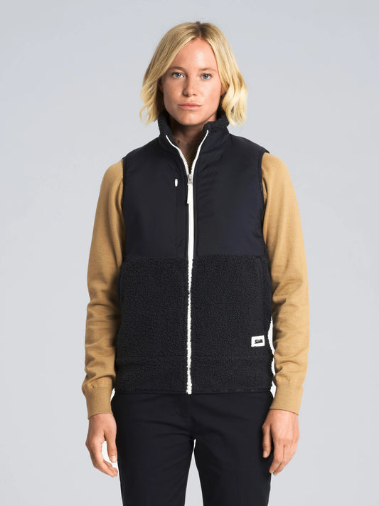 A person with short blond hair wears a tan long-sleeve shirt, black pants, and a SLAM ALTURA WS VEST in dark navy—featuring fleece and technical nylon construction with a white zipper—standing against a light gray background.