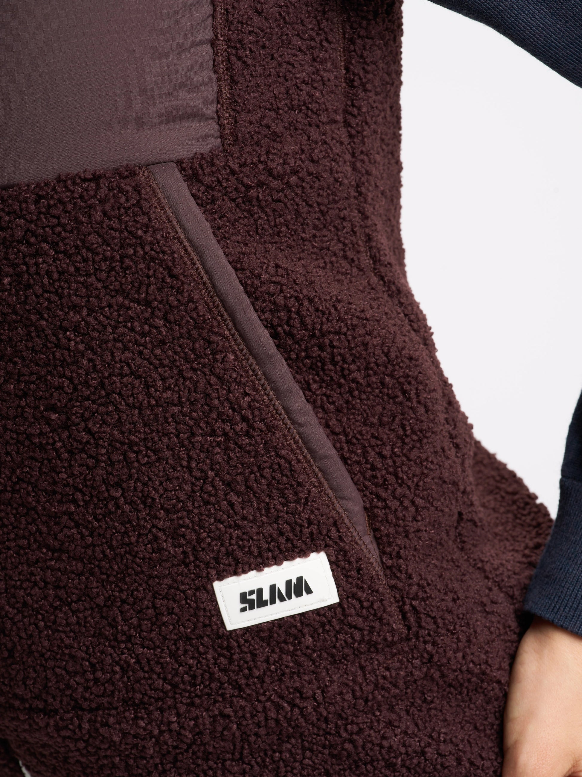 A close-up of a person wearing the ALTURA WS VEST by SLAM—a dark brown, textured fleece waistcoat featuring a diagonal zipper pocket and a rectangular white and black “SLAM” label. The person’s arm, in a dark sleeve, is partially visible.