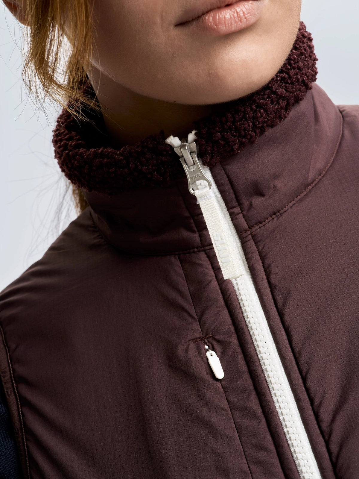 Close-up of a person wearing a brown zip-up jacket with a textured collar and white zipper, layered over the SLAM ALTURA WS VEST. The lower half of their face, along with part of their neck and hair, is visible.