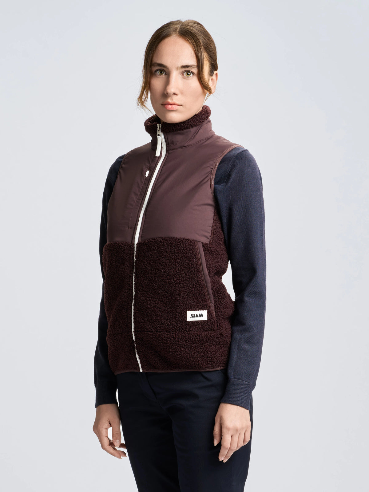 A woman stands facing forward, wearing the SLAM ALTURA WS VEST—a dark purple fleece waistcoat featuring a fleece lower half—layered over a long-sleeve navy shirt and dark pants. The background is plain and light-colored.