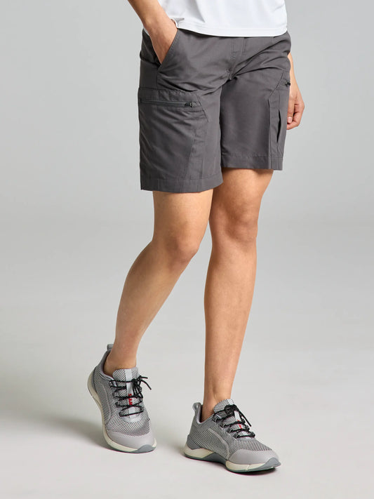 A person dressed in SLAM's ACTIVE WOMAN CARGO SHORTS in titanium, paired with a white shirt and gray sneakers, stands against a neutral gray background with hands in pockets. The shorts' practical, water-repellent design adds a stylish touch to this casual ensemble.