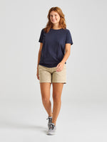 A person with shoulder-length hair is showcasing a summer style, wearing a navy T-shirt paired with SLAM's DECK WOMAN LIGHT CHINO SHORT in beige, and completed with gray sneakers. They are smiling against a plain, light-colored background.