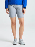 A person stands against a plain background wearing a blue jacket, SLAM WS TECH CHINO SHORT in light gray technical elastic fabric, and gray athletic shoes. Their hands are relaxed at their sides.