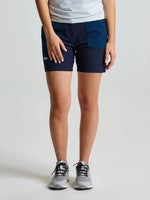 A person is wearing an OD WS CHINO SHORT by SLAM in DARK NAVY—durable, breathable shorts with side pockets made from Double Weave stretch fabric—paired with a white shirt and gray running shoes, standing against a plain light background.