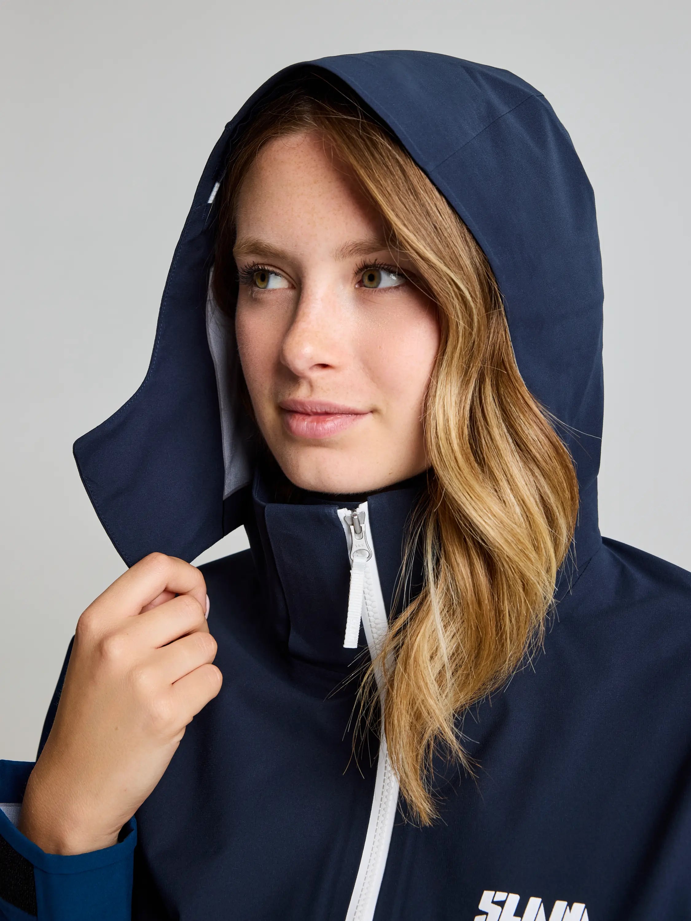 A person with long hair wears a SLAM FOIL WS HOODED JACKET, navy blue, breathable, featuring a white zipper and logo on the chest. The person adjusts the hood with one hand while looking to the side against a plain, light-colored background.