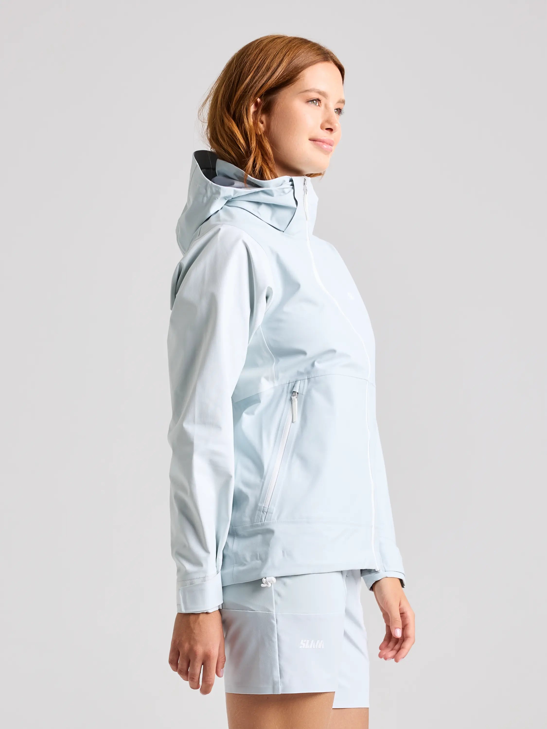 A person with shoulder-length hair, wearing SLAM's FOIL WS HOODED JACKET in light blue and matching shorts, stands in profile against a plain background. They smile slightly with their hands by their sides.