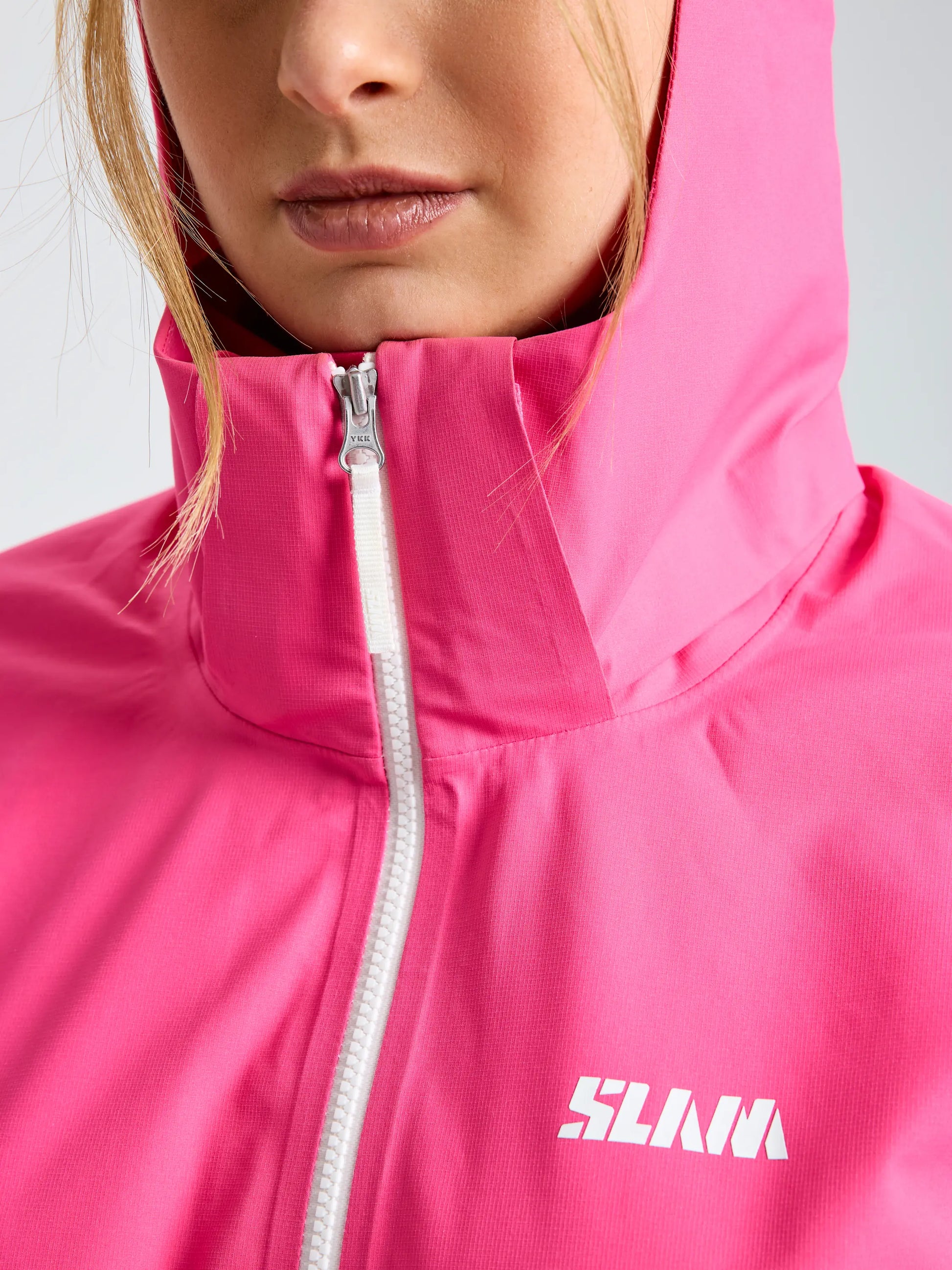A person wears the SLAM OD WS HOODED JACKET, a bright pink waterproof and breathable sailing jacket with a white zipper and "SLAM" on the chest. Their hair and lower face are visible, emphasizing the stylish design.