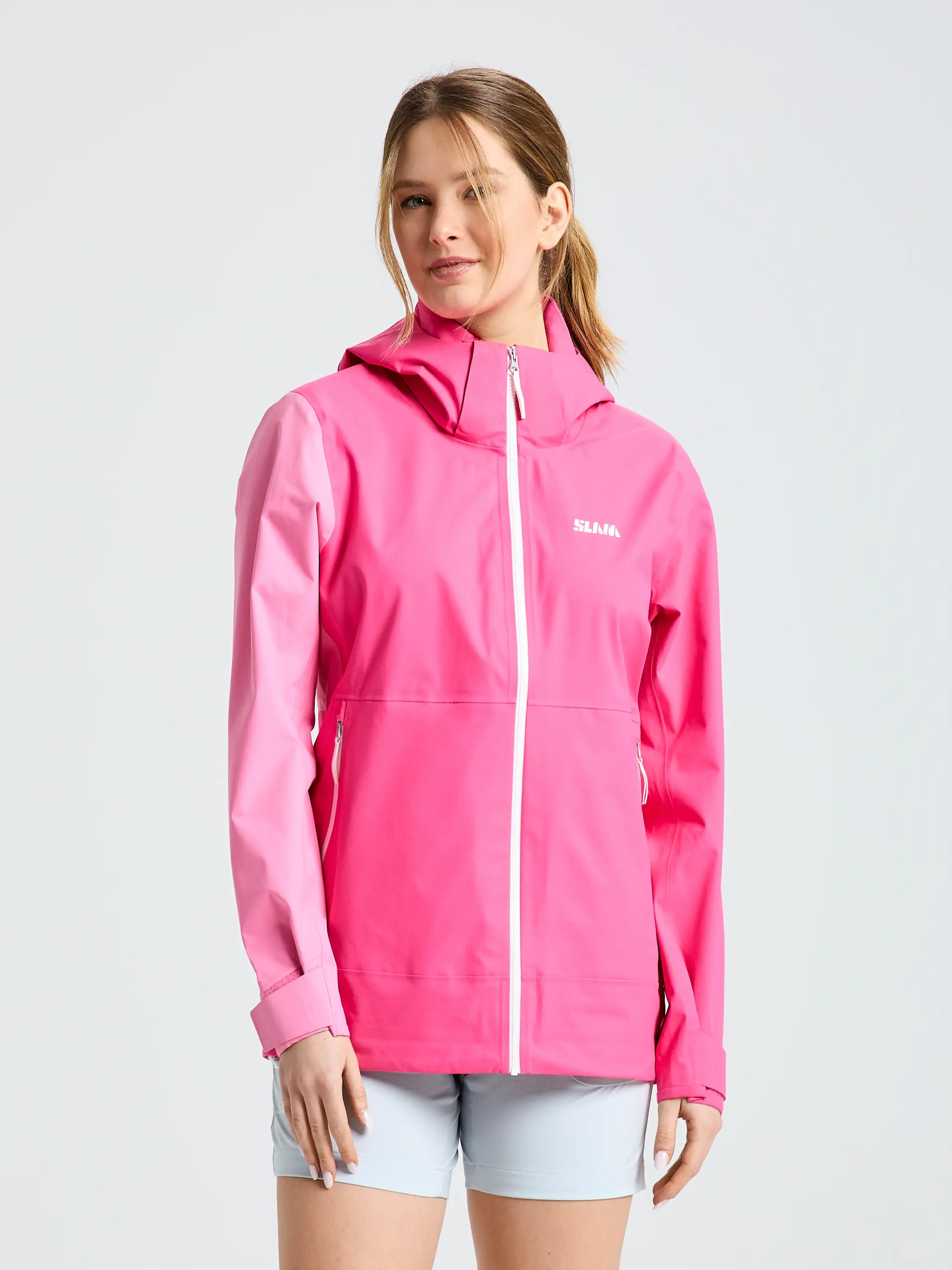 A woman wears the SLAM OD WS HOODED JACKET—bright pink, waterproof, breathable with a hood, white zipper and logo—paired with light gray shorts, standing against a plain light background.