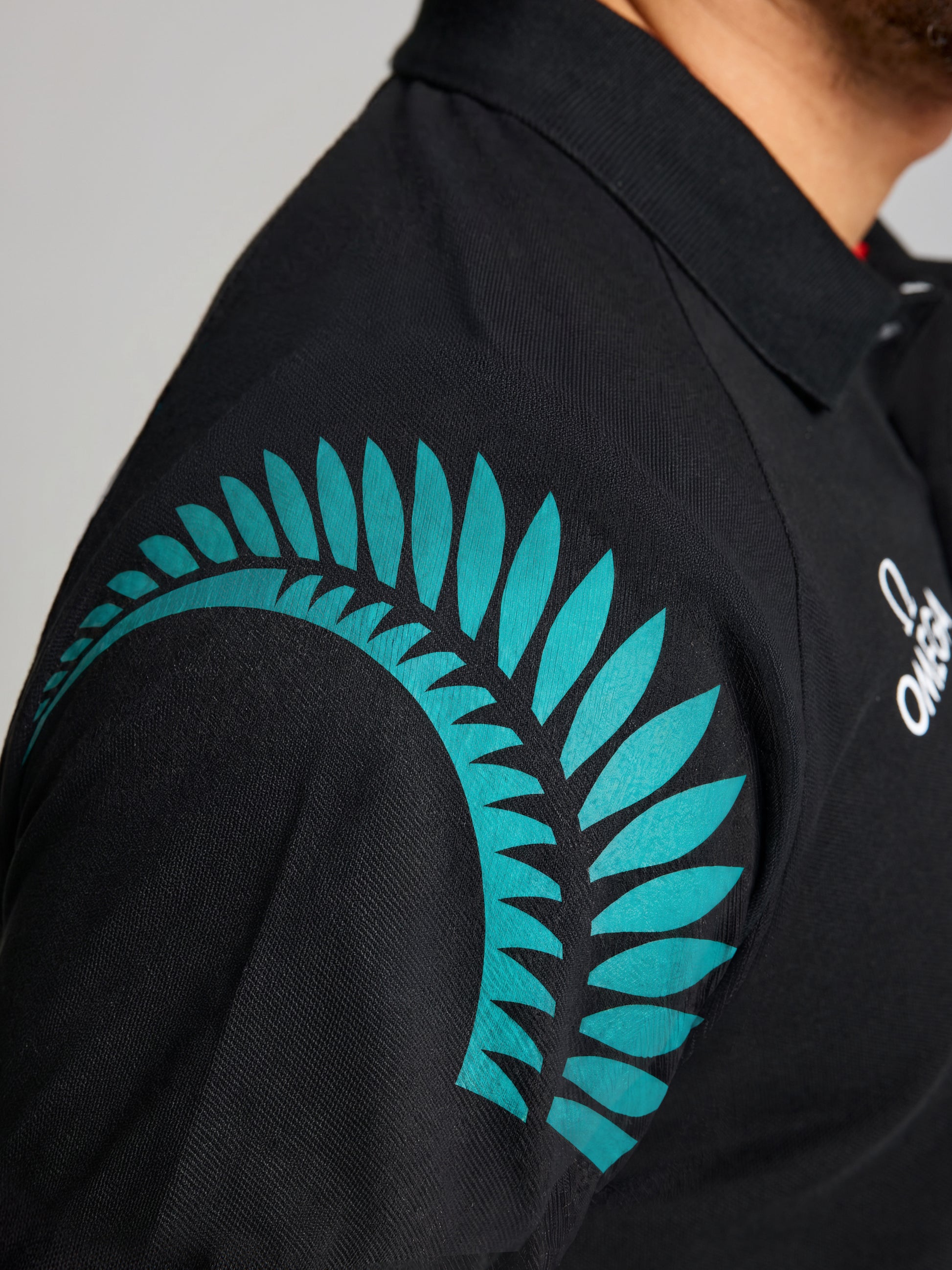 A close-up of a person wearing the SLAM ETNZ DECK POLO SS—a black men’s short-sleeve polo with a teal fern on the shoulder and a white chest logo. Only part of the individual's face and upper body is shown.