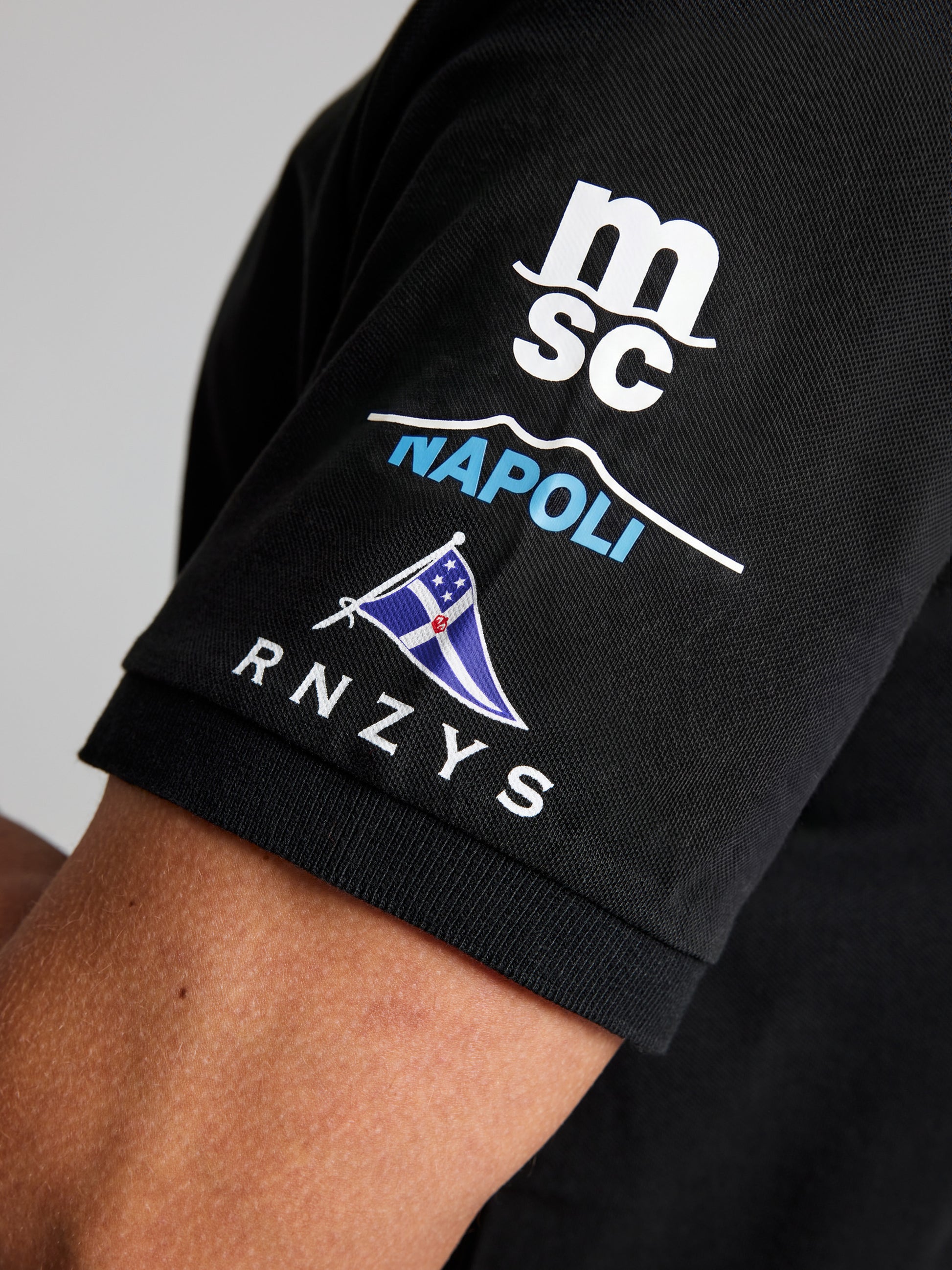 A close-up of the black SLAM ETNZ DECK POLO SS men's short-sleeve polo, inspired by Emirates Team New Zealand, featuring "msc," "NAPOLI," a flag graphic, and "RNZYS" logos printed in white and blue.