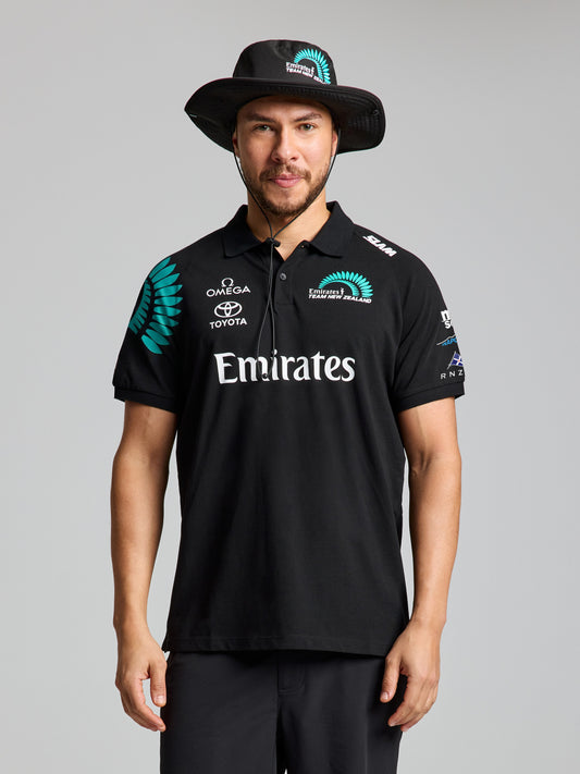 A man stands facing the camera against a plain light background, wearing the SLAM ETNZ DECK POLO SS in ETNZ BLACK with multiple sponsor logos, including Emirates, Omega, and Toyota, made from organic cotton.