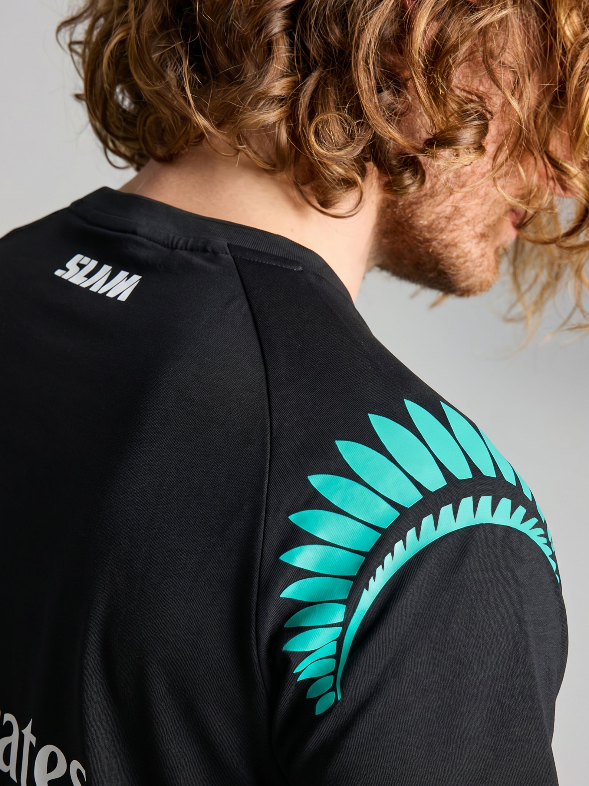 A close-up of a person with curly hair facing away, wearing the SLAM ETNZ DECK T-SHIRT, which features a teal feather design and white "SLAM" text on the back, inspired by Emirates Team New Zealand and the America's Cup.