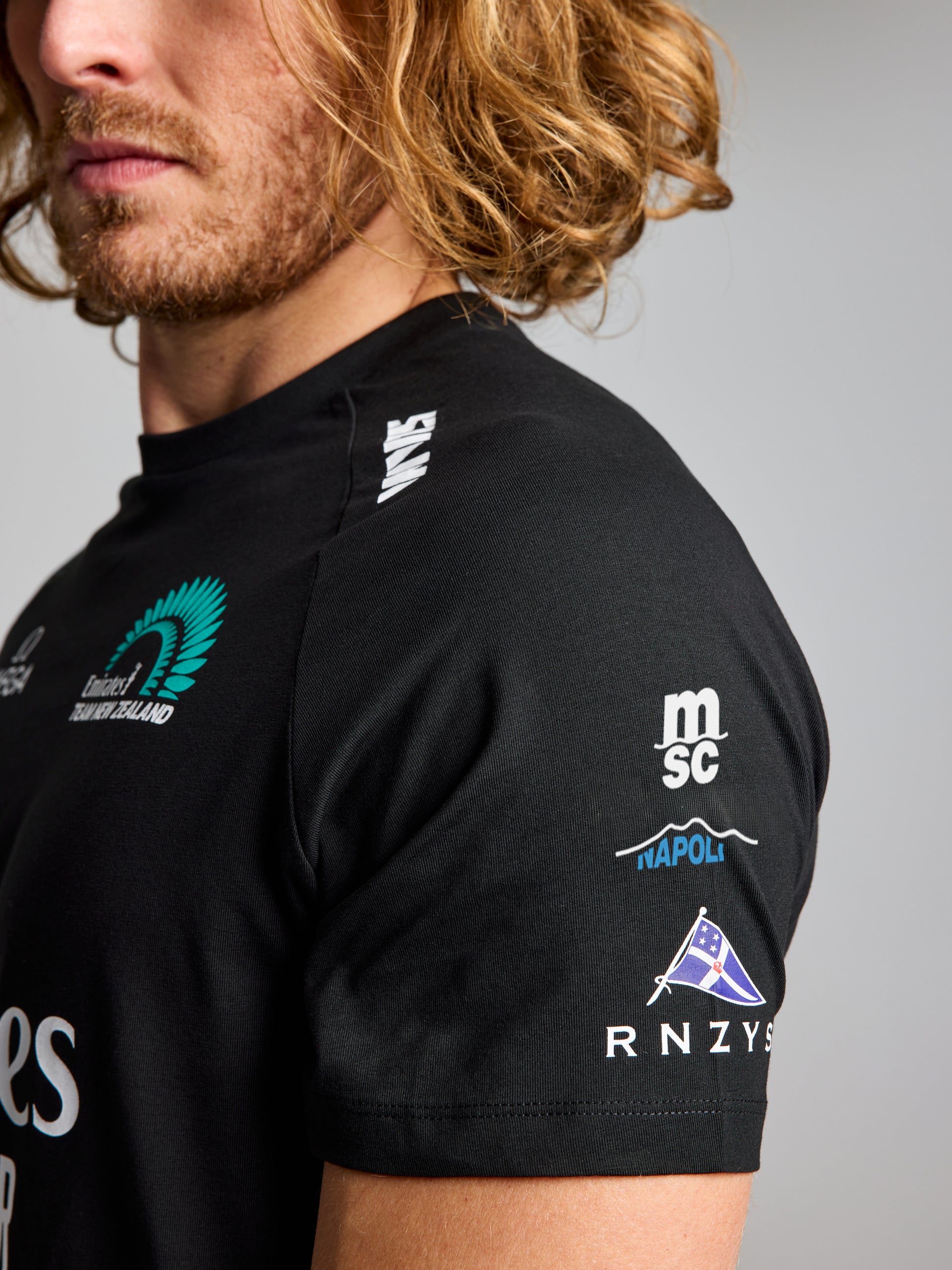 A person with long, wavy, light brown hair is shown from the shoulders up, wearing the SLAM ETNZ DECK T-SHIRT in black with white and green Emirates Team New Zealand logos on the shoulder and sleeve. Face is partially out of frame.