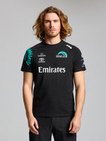 A man with long, wavy hair faces forward wearing the SLAM ETNZ DECK T-SHIRT in ETNZ BLACK, featuring Omega, Toyota, and Emirates logos. The plain light gray background highlights his link to the America’s Cup team.