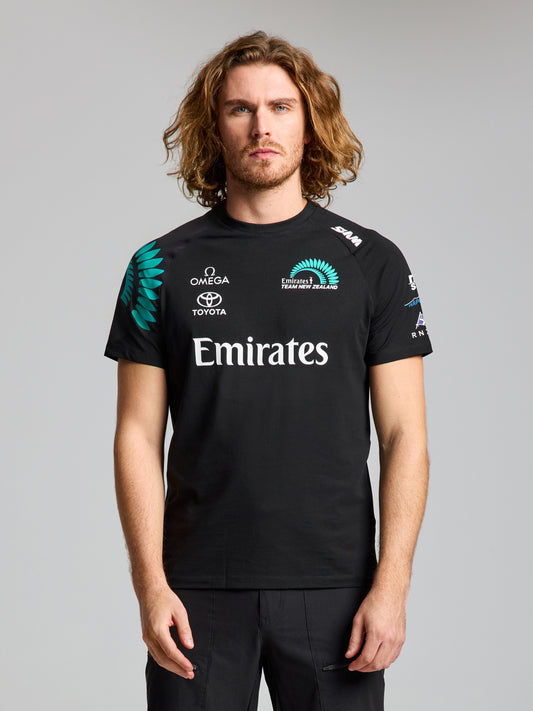 A man with long, wavy hair faces forward wearing the SLAM ETNZ DECK T-SHIRT in ETNZ BLACK, featuring Omega, Toyota, and Emirates logos. The plain light gray background highlights his link to the America’s Cup team.