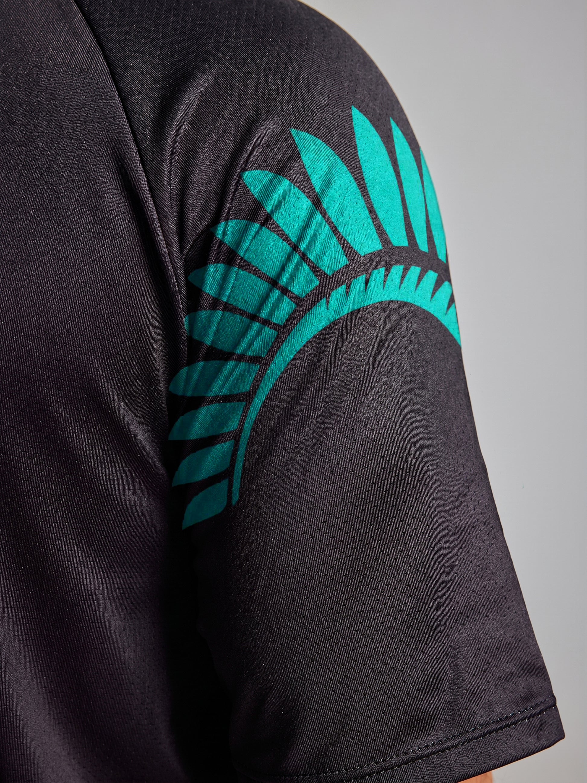 Close-up of the SLAM ETNZ SUNBLOCK SS: a black short-sleeve t-shirt with a teal feather-like graphic on the shoulder. Features UV-protective fabric inspired by Emirates Team New Zealand. Neutral background.