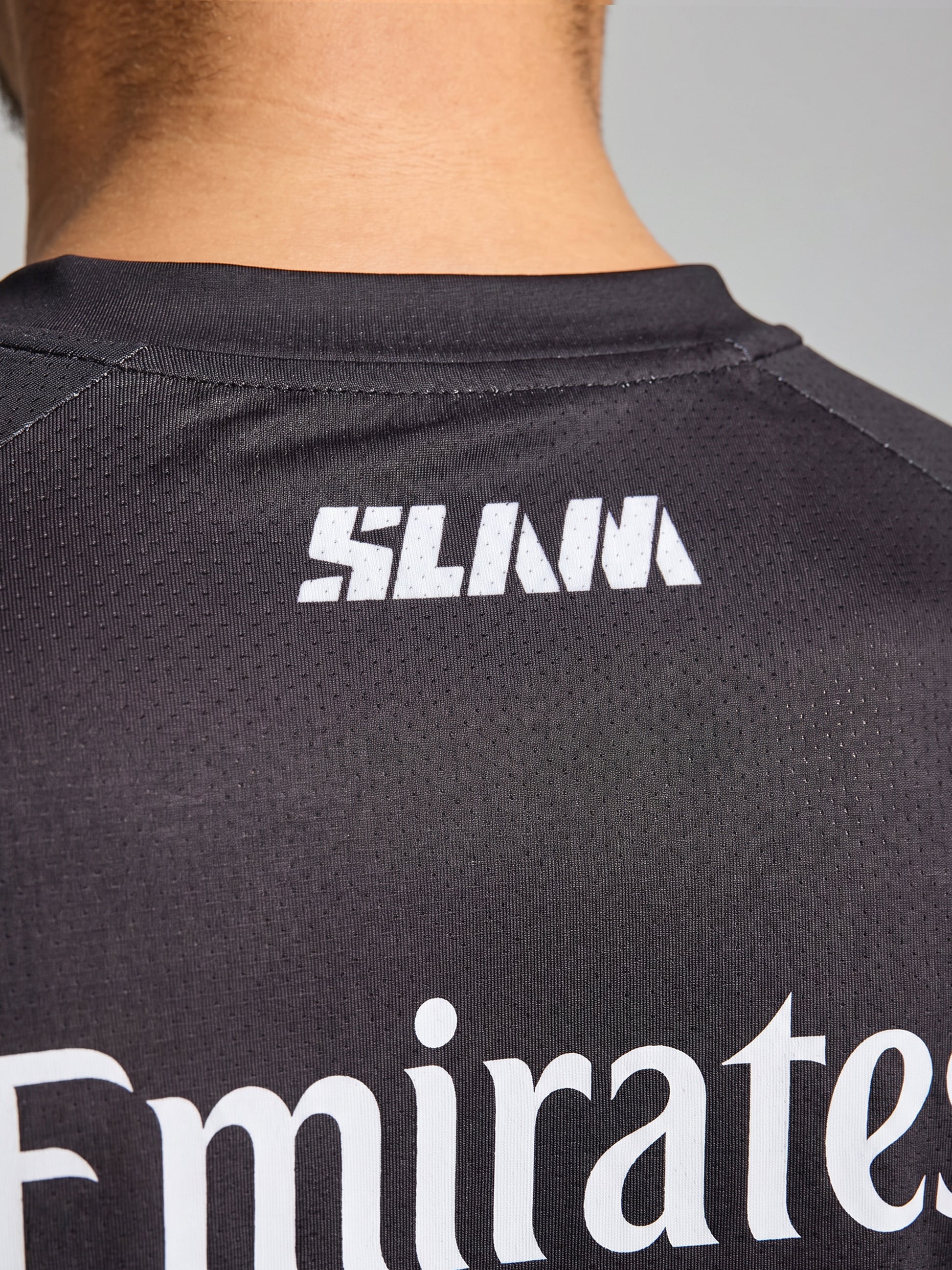 Close-up of the upper back of a person wearing a SLAM ETNZ SUNBLOCK SS T-shirt, made with UV-protective fabric. "SLANA" is printed in white near the neck, with part of "Emirates" visible lower down.