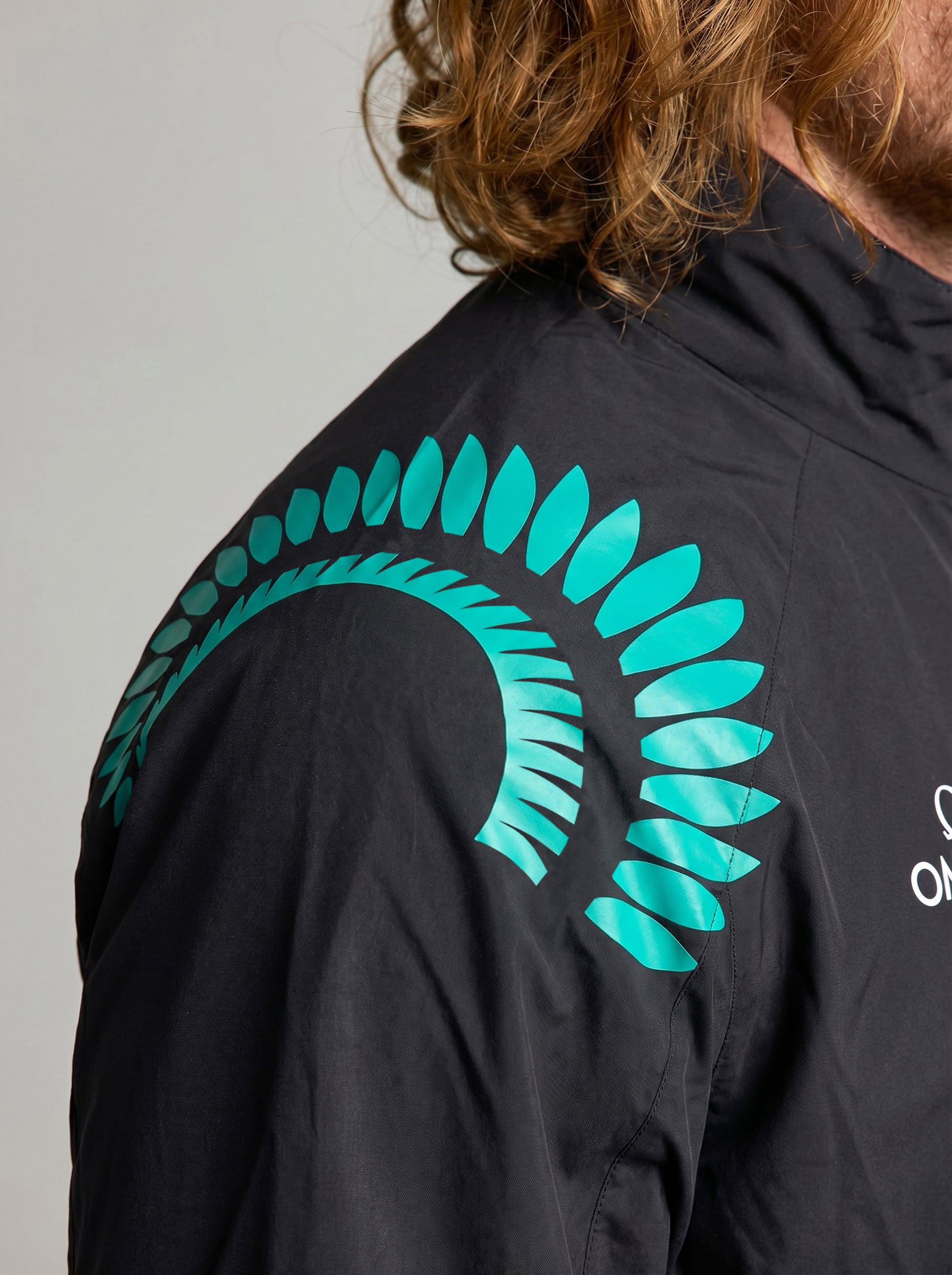 A person with long, wavy hair wears a black SLAM ETNZ DECK WP LGT JACKET with a teal fern on the shoulder. Only the upper right torso and part of the head are visible.