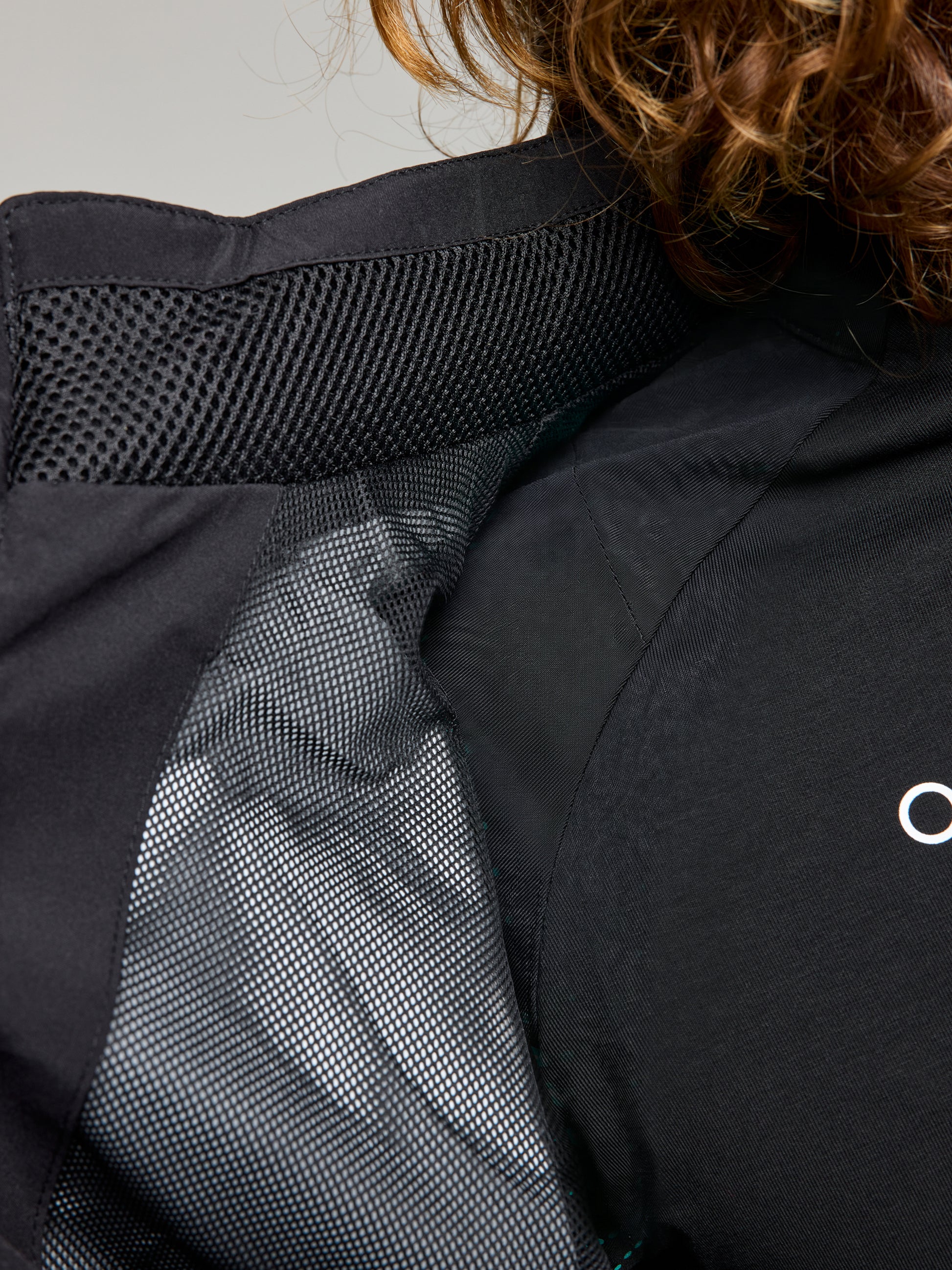 Close-up of a shoulder wearing the SLAM ETNZ DECK WP LGT JACKET, a black waterproof summer jacket with mesh ventilation. Detailed stitching and fabric texture are visible, with brown, curly hair partly in the top right corner.