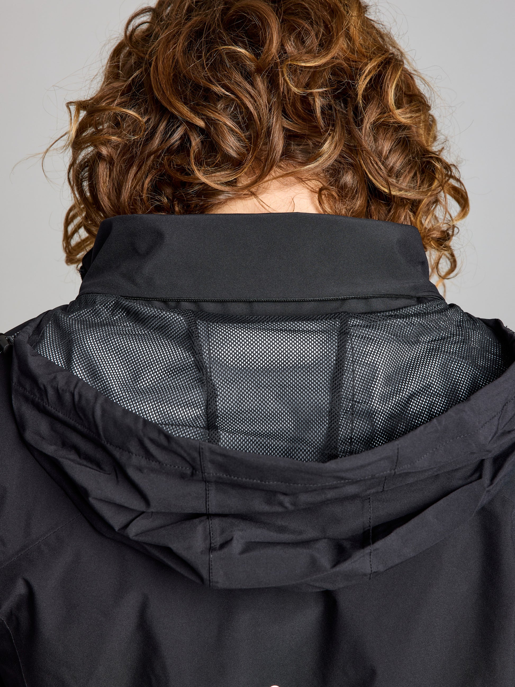 A person with curly brown hair is shown from behind, wearing a SLAM ETNZ DECK WP LGT JACKET with a partially open collar revealing a gray mesh lining. The waterproof summer jacket stands out against the plain background.