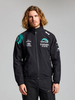 A person with long wavy hair wears the SLAM ETNZ DECK WP LGT JACKET, a waterproof summer jacket with Emirates Team New Zealand, Omega, and Toyota sponsor logos, standing against a plain gray background and looking at the camera.