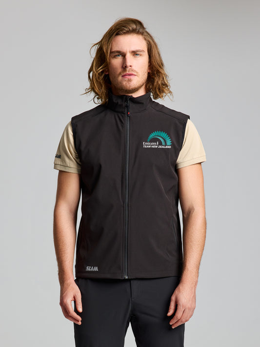 A man with shoulder-length hair wears a beige short-sleeve shirt and the SLAM ETNZ ACT SOFTSHELL VEST in BLACK INK, featuring an embroidered logo and "Environment – Your New Zealand" text, standing against a plain gray background.