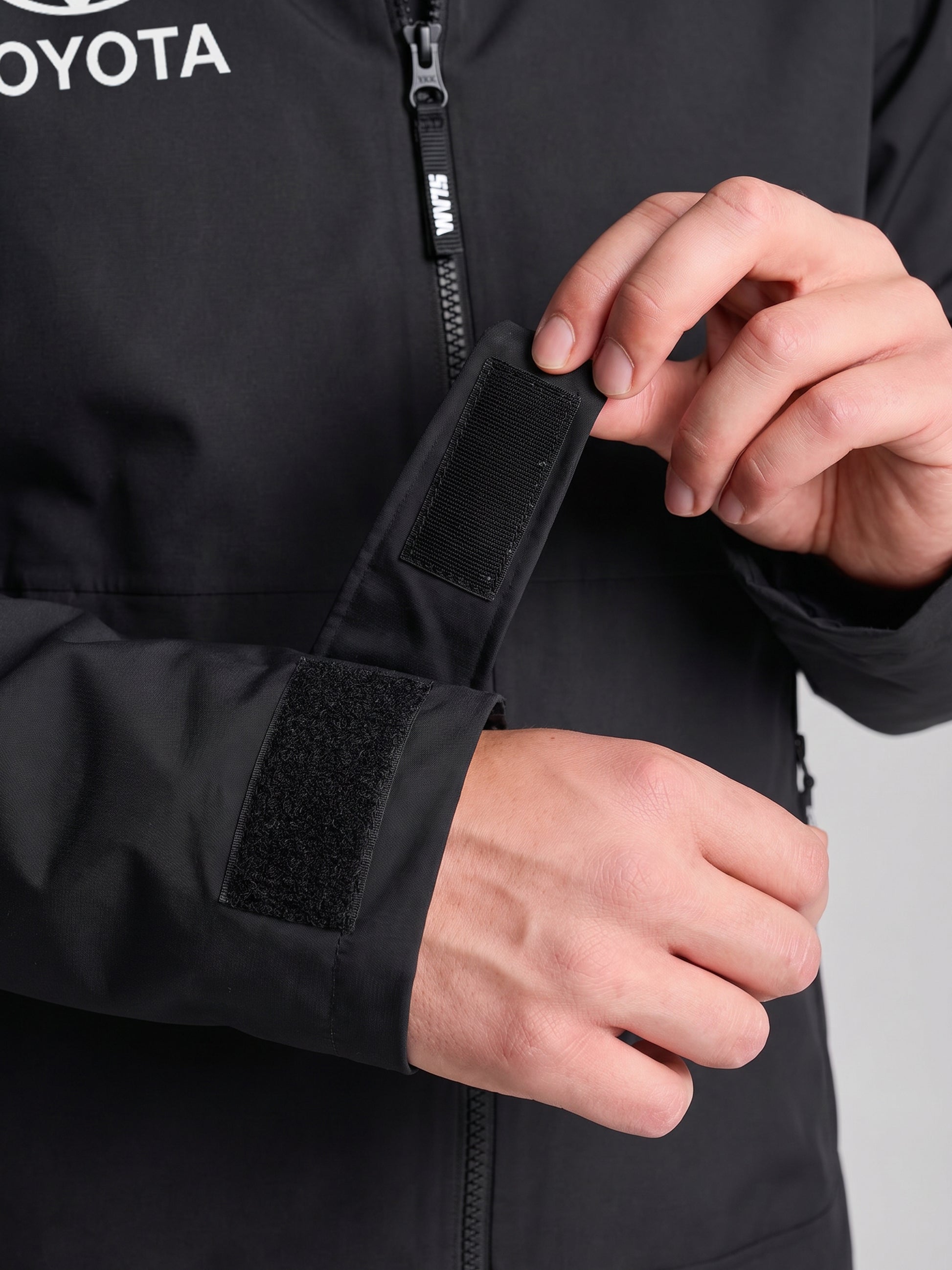 A person adjusts the Velcro cuff strap on a black SLAM ETNZ OD HOODED JACKET. The jacket features a zipper and visible "TOYOTA" logo, marking it as official Emirates Team New Zealand replica gear for the Americas Cup.