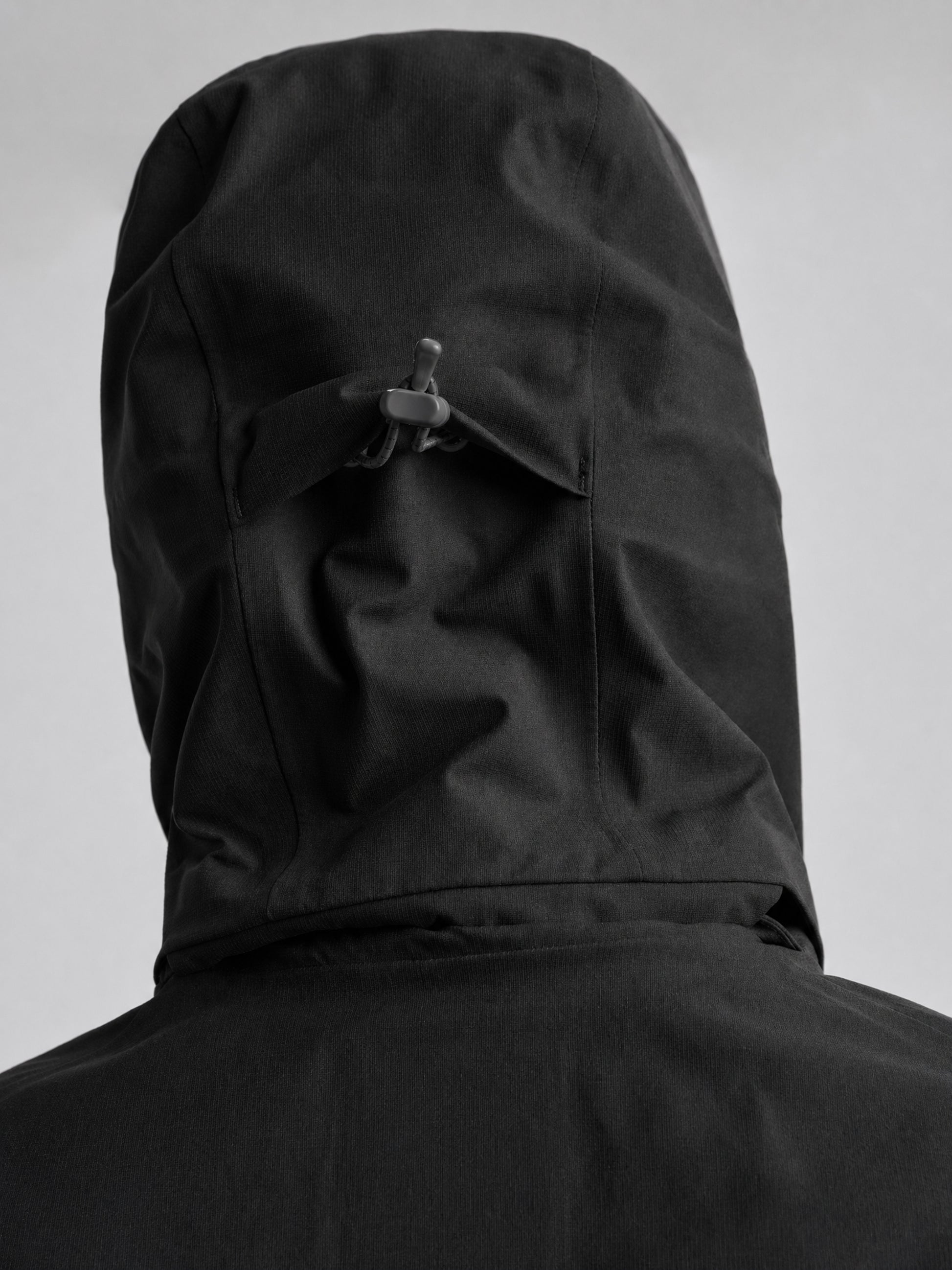 A person, seen from behind, wears a SLAM ETNZ OD HOODED JACKET inspired by the Emirates Team New Zealand Replica. The hood is up with an adjustable toggle at the back. The background is plain and light-colored.