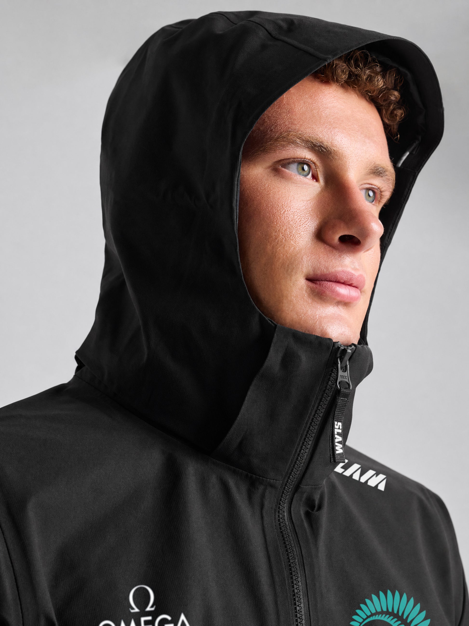 A person with light skin and curly hair wears the SLAM ETNZ OD HOODED JACKET, featuring white Emirates Team New Zealand Replica logos on the chest and shoulder, standing against a plain gray background.