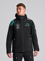 A person stands slightly to the side, wearing a SLAM ETNZ OD HOODED JACKET in ETNZ BLACK, an Emirates Team New Zealand replica with sponsor logos and green wing-like designs, set against a plain light grey background.