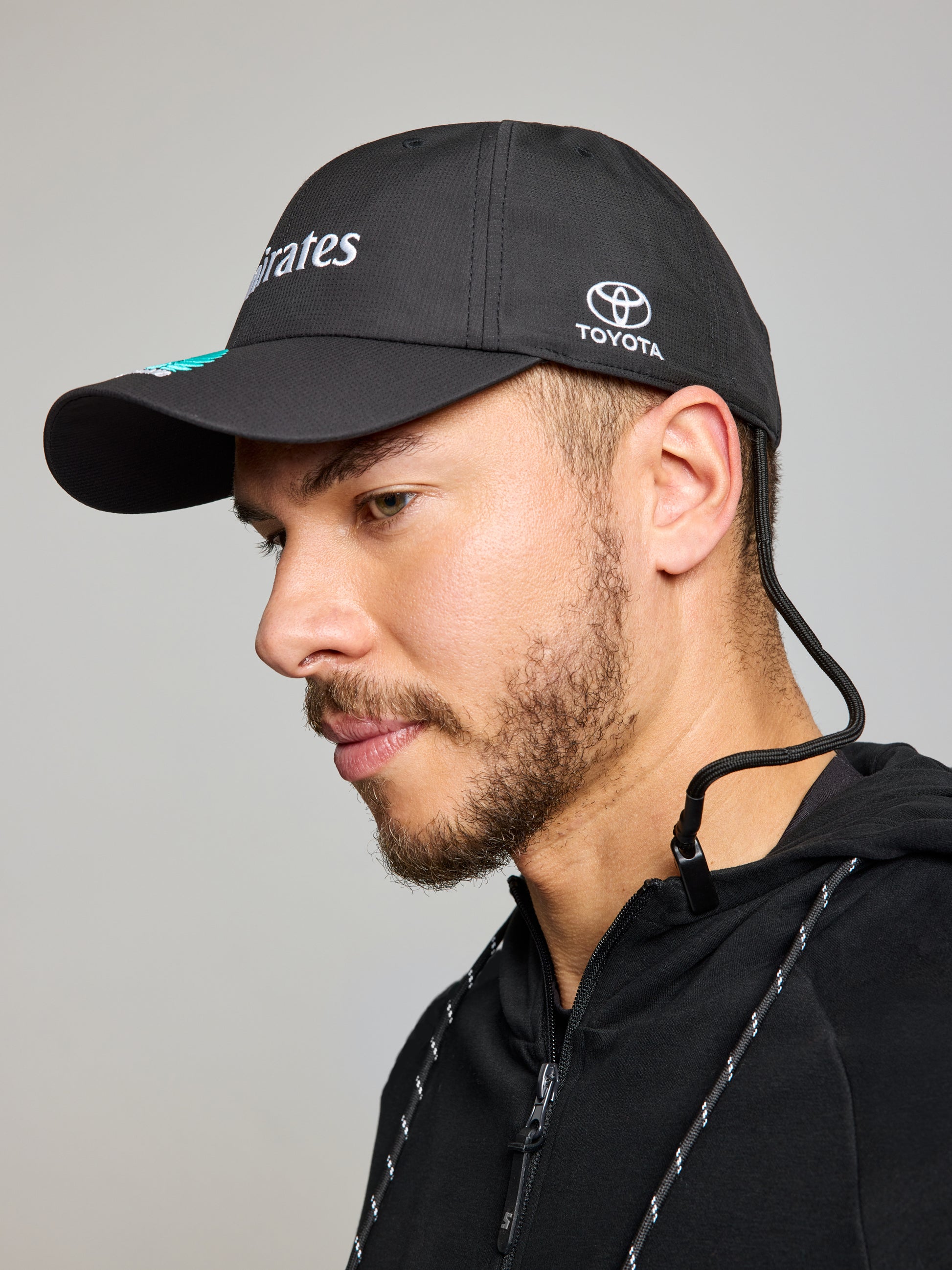 A man with a trimmed beard wears a black SLAM ETNZ TECH CAP with "Toyota" and "Emirates" logos, plus a black zip-up hoodie. Facing left against a plain light background, he showcases Emirates Team New Zealand and the America’s Cup spirit.