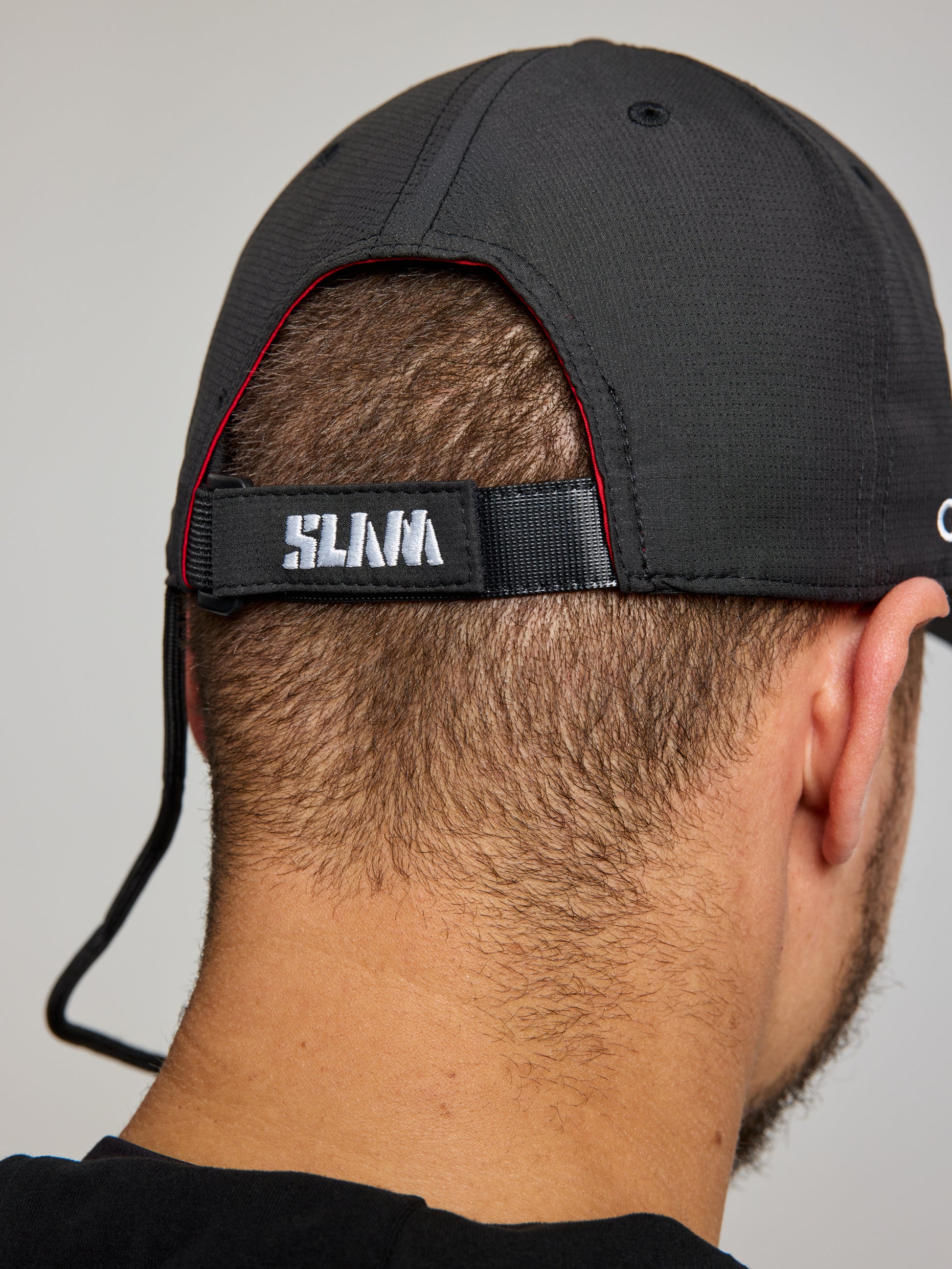 A close-up shows the back of a person’s head wearing an ETNZ TECH CAP by SLAM, inspired by Emirates Team New Zealand and the America’s Cup, with "SLNA" embroidered in white on the adjustable strap. The person's short hair and ear are visible.