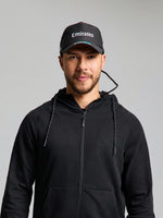 A person wearing a black zip-up hoodie and a SLAM ETNZ TECH CAP in ETNZ BLACK stands against a plain light gray background, facing the camera with a neutral expression.
