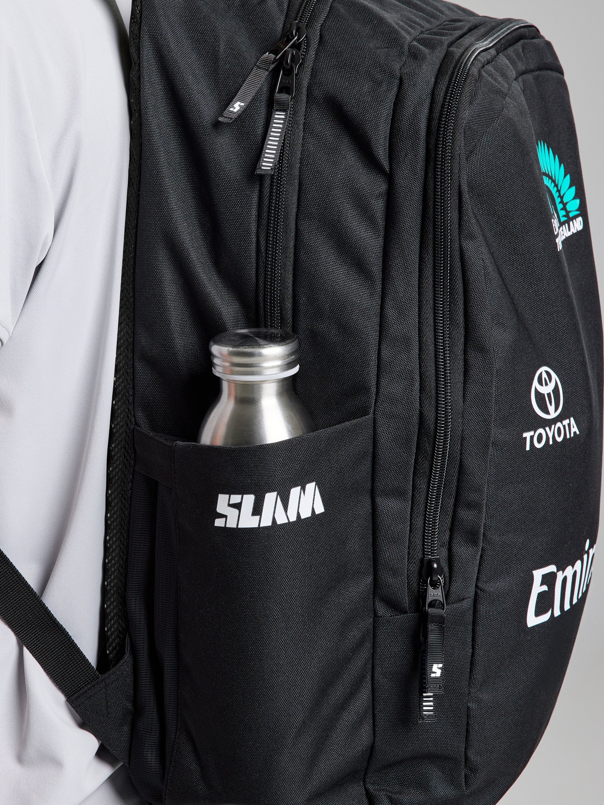 A close-up of the SLAM ETNZ BACKPACK shows multiple zippered pockets, a silver water bottle in a side pocket, and white logos such as SLAM, Toyota, Emirates Team New Zealand, and America's Cup on the front.