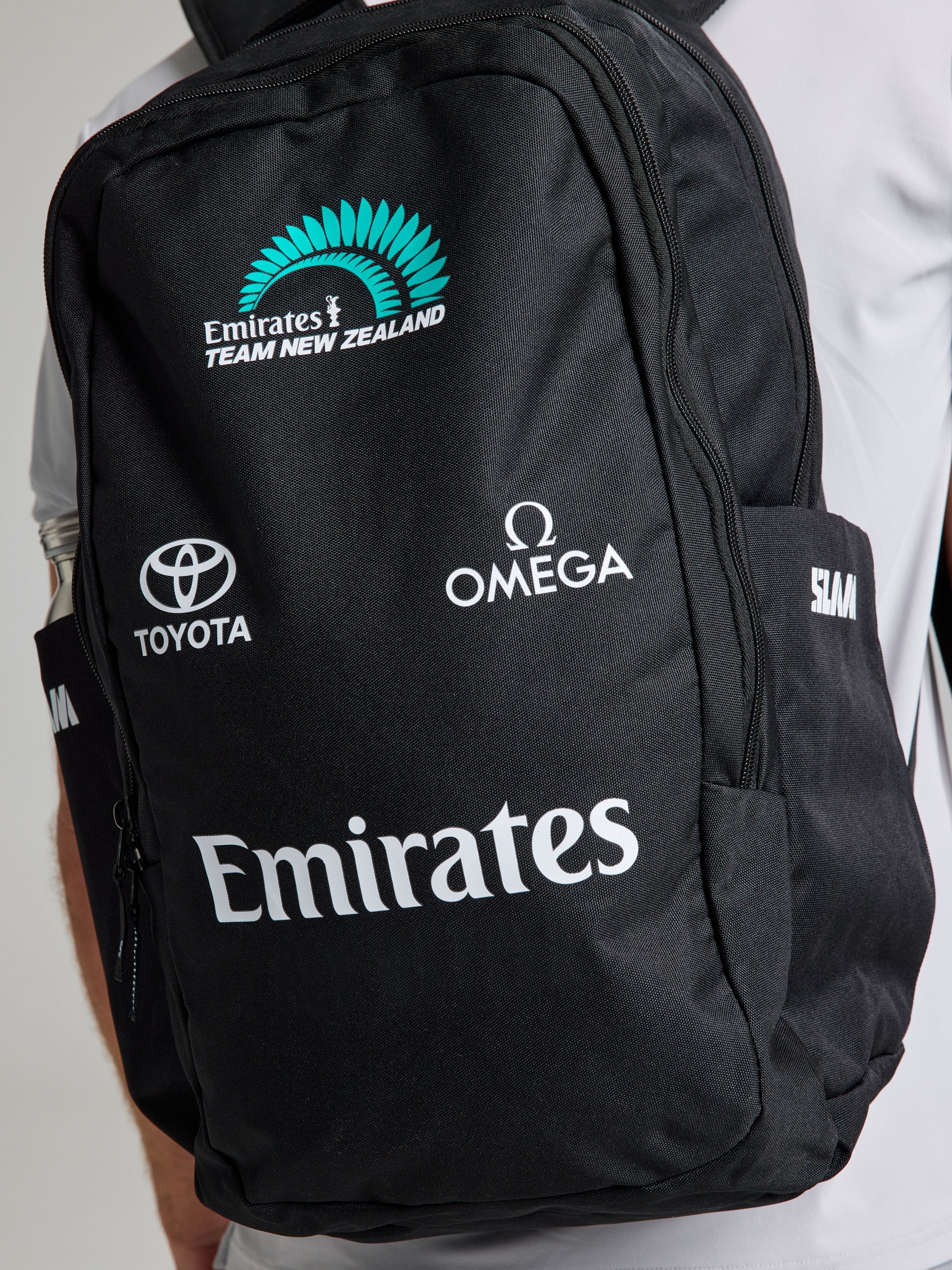 A person wearing an SLAM ETNZ BACKPACK with Emirates Team New Zealand, Toyota, Omega, and Emirates logos, linked to the America's Cup, over a light-colored shirt.