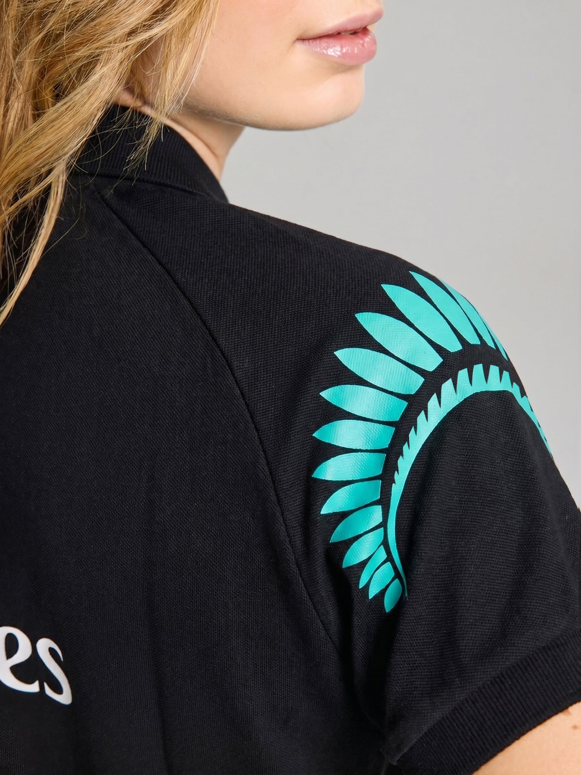 A close-up shows a person wearing the SLAM ETNZ DECK WS POLO SS, a black polo featuring a turquoise geometric design on the sleeve. Only the lower face and part of the shoulder are visible.