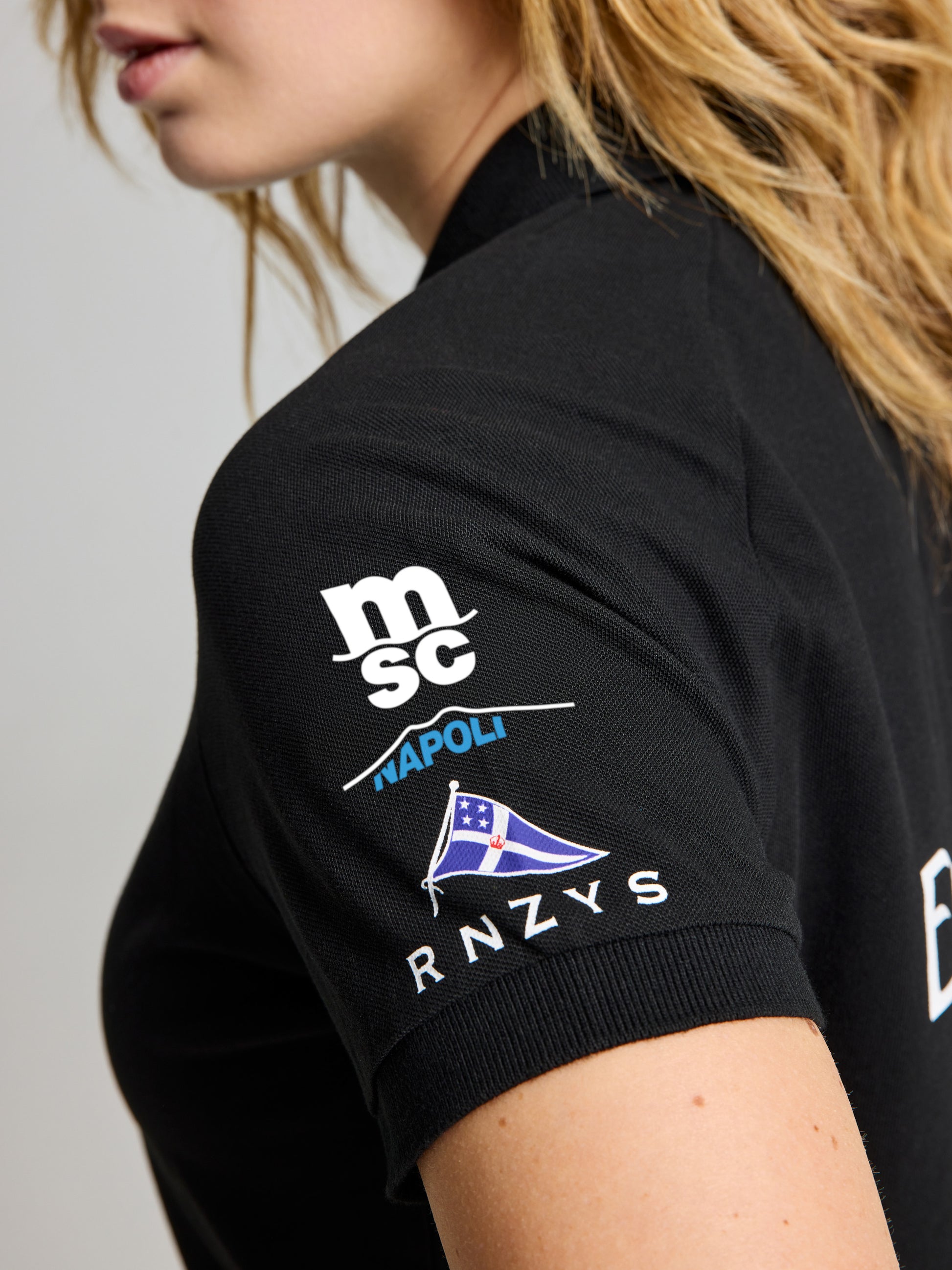 A close-up of a woman's upper arm and shoulder wearing the black SLAM ETNZ DECK WS POLO SS with white logos, including "msc," "NAPOLI," a flag graphic, and "RNZYS." Her face is partially visible.