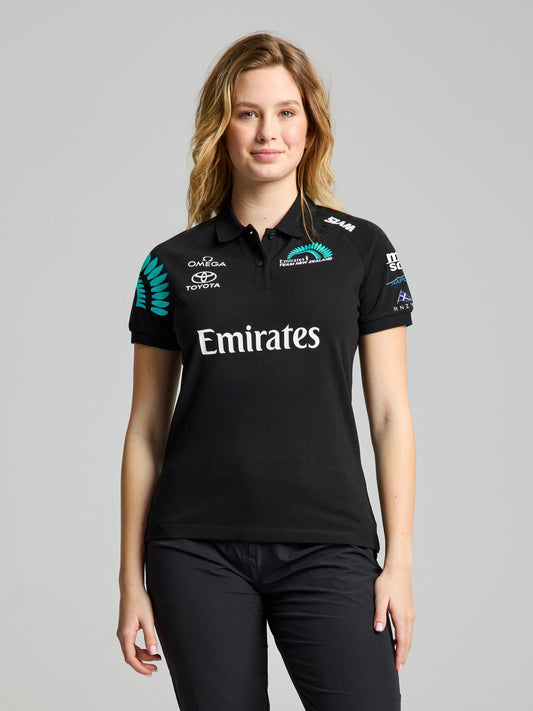 A woman with light brown hair faces forward wearing the SLAM ETNZ DECK WS POLO SS in ETNZ BLACK, featuring sponsor logos and black pants, set against a plain gray background.